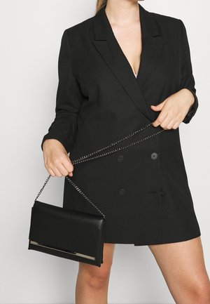 Nainen, joka pukeutunut mustaan kaksiriviseen bleiserimekkoon ja pitää kädessään pientä mustaa clutch-laukkua, jossa on hopeinen ketjuhihna.