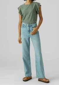 Haut vert à manches courtes avec des détails en œillet, associé à un jean flare bleu clair. Porté avec des sandales à plateforme marron.