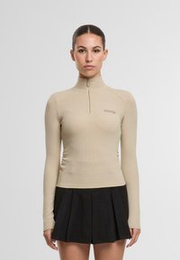 Bežinė ilgomis rankovėmis palaidinė su aukštu kaklu ir pusiau užtrauktuku. Tekstūruotas audinys su prigludusia siluetu. Juoda klostuota sijonas dėvima žemiau.