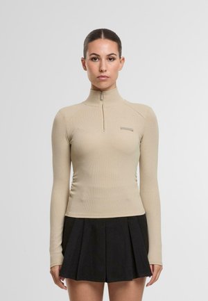 Beżowa bluzka z długim rękawem, z wysokim kołnierzem i pół zamkiem. Teksturowany materiał o dopasowanej sylwetce. Czarna spódnica plisowana noszona poniżej.