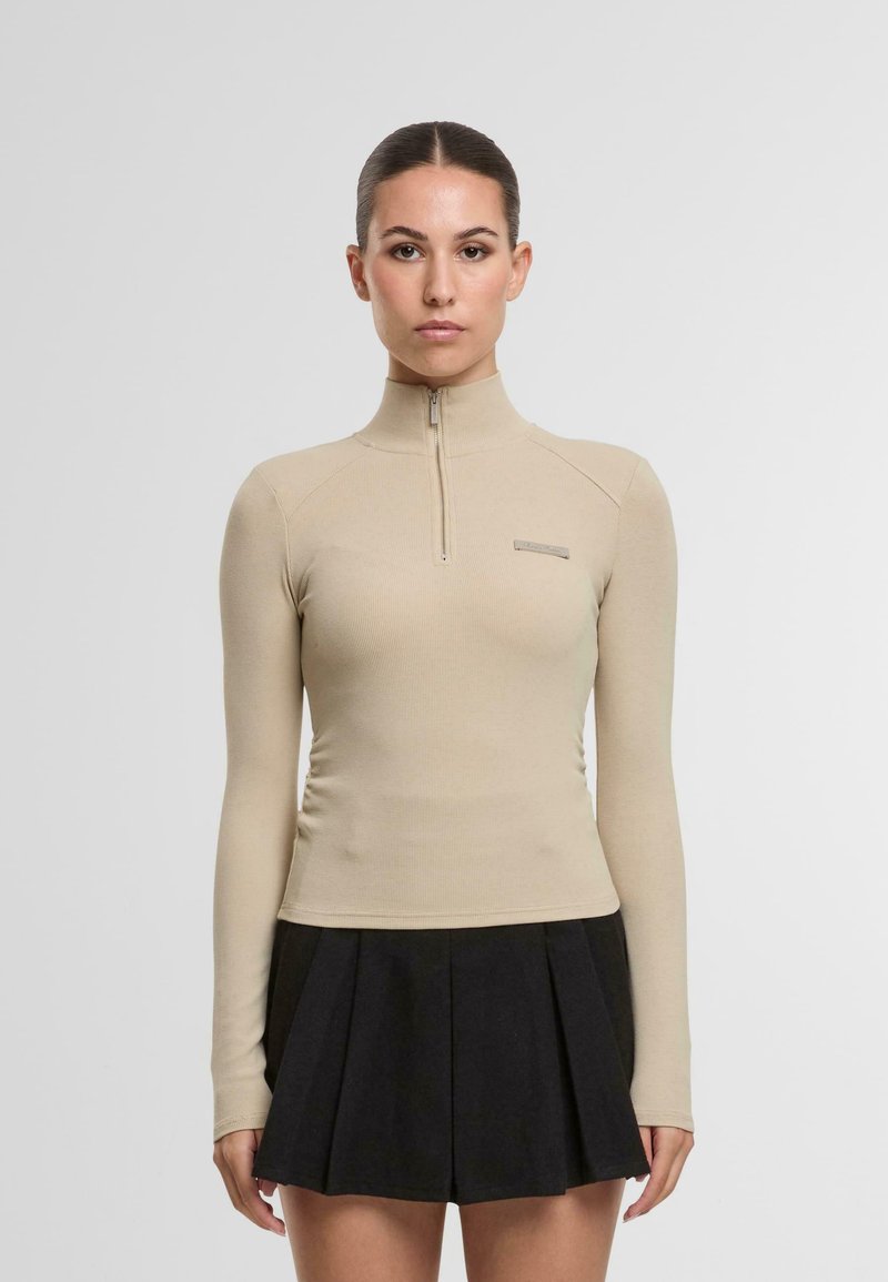 Bežinė ilgomis rankovėmis palaidinė su aukštu kaklu ir pusiau užtrauktuku. Tekstūruotas audinys su prigludusia siluetu. Juoda klostuota sijonas dėvima žemiau.