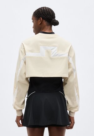 Donna con i capelli intrecciati vista di spalle, indossa una giacca beige corta con motivo riflettente e una gonna nera con piping bianco.