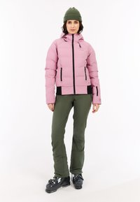 Světle růžová péřová bunda s kapucí, černými akcenty a zipy, spárovaná s olivově zelenými sněhovými kalhotami a černými lyžařskými botami.