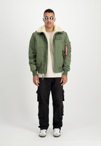 Veste bomber verte avec col en fausse fourrure, fermeture éclair à l'avant et patch ; portée par-dessus une chemise beige et associée à un pantalon cargo noir et des baskets blanches.