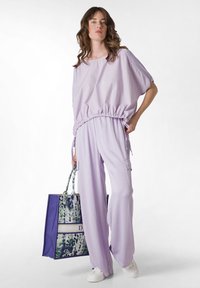 Lavendel, lös passform topp och byxor med elastisk midjebandsdetalj; textil med struktur. Modellen håller en flerfärgad mönstrad tote-väsk och bär vita sneakers.