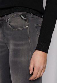 Vaqueros de mezclilla gris con una textura desgastada, que presentan una etiqueta negra con "REPLAY" en el bolsillo trasero y detalles en hardware plateado.