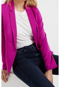 Blazer fuchsia au design structuré, avec des revers en pointe et un tissu lisse, associé à un t-shirt blanc et un jean foncé.