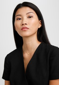 Collier pendentif en cœur doré sur une fine chaîne, porté sur un haut noir à décolleté en V profond, avec des manches courtes et une texture de tissu lisse.