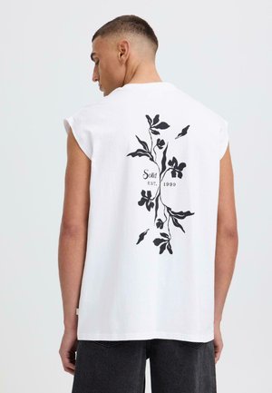 Homme portant un débardeur blanc avec un motif floral noir et le texte "Solid EST 1999" au dos, associé à un jean foncé.