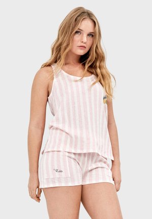 Joven con cabello largo y rubio que lleva un top sin mangas a rayas verticales rosas y blancas y pantalones cortos a juego con un pequeño detalle de piña.