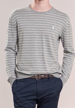 Man met handen in de zakken, die een grijs-wit gestreept shirt met lange mouwen en een donkerblauwe broek met een gevlochten bruine riem draagt.