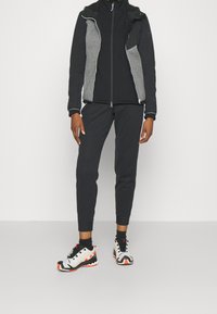 Veste de sport noire avec des panneaux gris, fermeture éclair à l'avant ; assortie à un pantalon de jogging noir et des chaussures de course multicolores avec des accents orange.