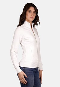 Giacca bianca con zip, colletto alto, vestibilità slim e polsini a costine. Presenta una zip dorata e un dettaglio con logo sul petto.
