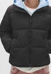 Personne portant une veste matelassée noire avec les mains dans les poches, une capuche bleu clair, une chemise blanche en dessous, et un jean rose délavé.