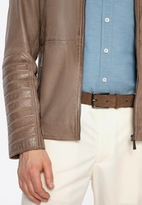 Giubbotto di pelle marrone con maniche testurizzate, chiusura con zip e dettagli a coste, abbinato a una camicia azzurra chiara e una cintura marrone. Pantaloni bianchi.