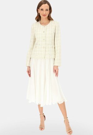 Kobieta nosząca jasną, teksturowaną zapinaną na guziki marynarkę z kieszeniami oraz zwiewną białą spódnicę midi, pozująca w beżowych szpilkach z paskiem wokół kostki.