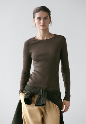 Vrouw met een bruine geribde top met lange mouwen, beige broek en een donker jasje om de taille gestrikt, staand tegen een effen lichte achtergrond.