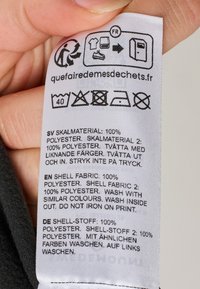 Etichetta di cura con istruzioni per la manutenzione e dettagli sui materiali; tessuto bianco con testo nero. Istruzioni in inglese, svedese e tedesco.