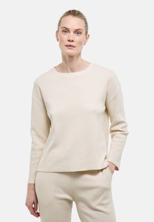 Femme aux cheveux blonds portant un haut à manches longues beige et un pantalon assorti, debout avec une main dans la poche, sur un fond blanc uni.