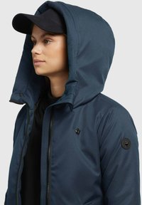 Marineblaues Kapuzenjacke mit glatter Textur, ausgestattet mit einem Reißverschluss vorne, schwarzen Beschlägen und einem kleinen Logo auf der Brust und dem Ärmel.