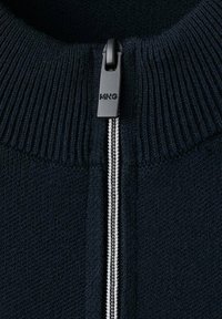 Makro fotografija crnog pletenog odjevnog predmeta sa srebrnim patentnim zatvaračem i crnim privjeskom patentnog zatvarača s oznakom "MNG" na rebrastom ovratniku.