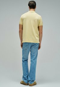 Hombre con camisa polo de manga corta de color amarillo pálido, jeans azules y zapatos color marrón claro, de pie de espaldas contra un fondo liso.