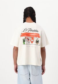 Cream-colored cotton t-shirt with short sleeves, featuring a colorful bistro illustration and "Le Parisien BISTRO" text on the back.