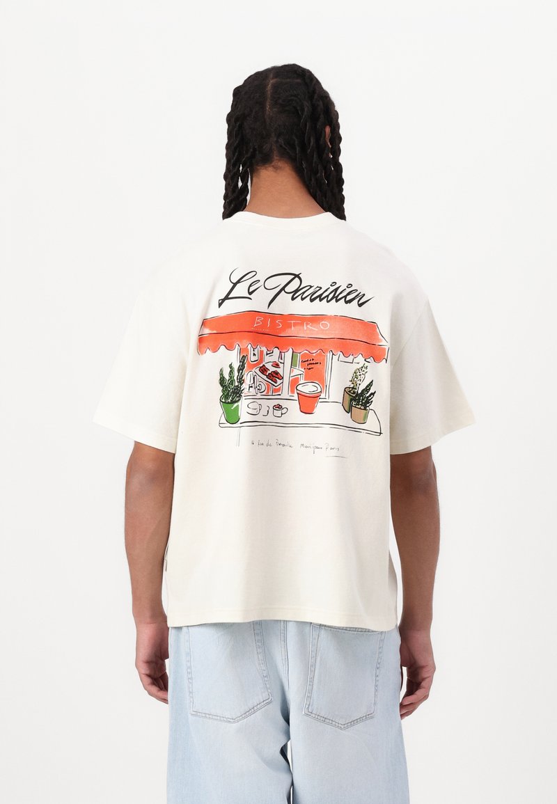 Cream-colored cotton t-shirt with short sleeves, featuring a colorful bistro illustration and "Le Parisien BISTRO" text on the back.