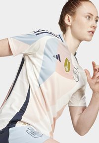 Jersey atlético blanco con formas geométricas en tonos pastel de azul, rosa y crema, con rayas negras y emblemas en el pecho y la manga.