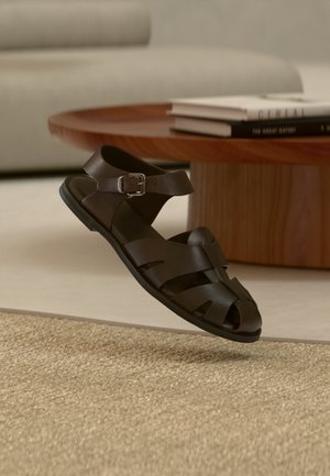 Mørkebrun lær herresandal med spenne, hvilende på beige teppe i en minimalistisk stue med trebord og bøker.