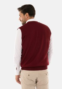 Gilet a maglia bordeaux con texture a coste, indossato sopra una camicia bianca a maniche lunghe. Pantaloni beige con tasche posteriori con bottoni.