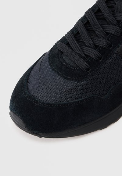 Εγγύς λήψη του μπροστινού τμήματος ενός μαύρου αθλητικού παπουτσιού που δείχνει υφασμάτινη υφή, επενδύσεις από σουέτ και σφιχτά δεμένα μαύρα κορδόνια σε λευκό φόντο.