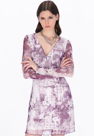 Giovane donna con lunghi capelli castani, che indossa un vestito a maniche lunghe con motivo viola e bianco e collane a catena d'argento sovrapposte, con le braccia incrociate.