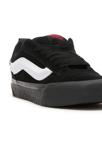 Sneaker in suede nera con un design a onda bianca, dotato di suola in gomma nera e una striscia grigia come accentuazione. Etichetta rossa con logo sulla linguetta.