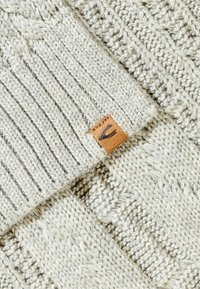 Pull en maille de couleur gris clair, avec une texture côtelée et une étiquette en cuir portant un logo. Le tissu présente un design en maille douce et épaisse.