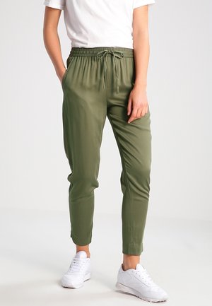 Person trägt olivgrüne Kordelzughose, weiße Sneaker und ein weißes Kurzarmhemd, steht mit einer Hand in der Tasche.