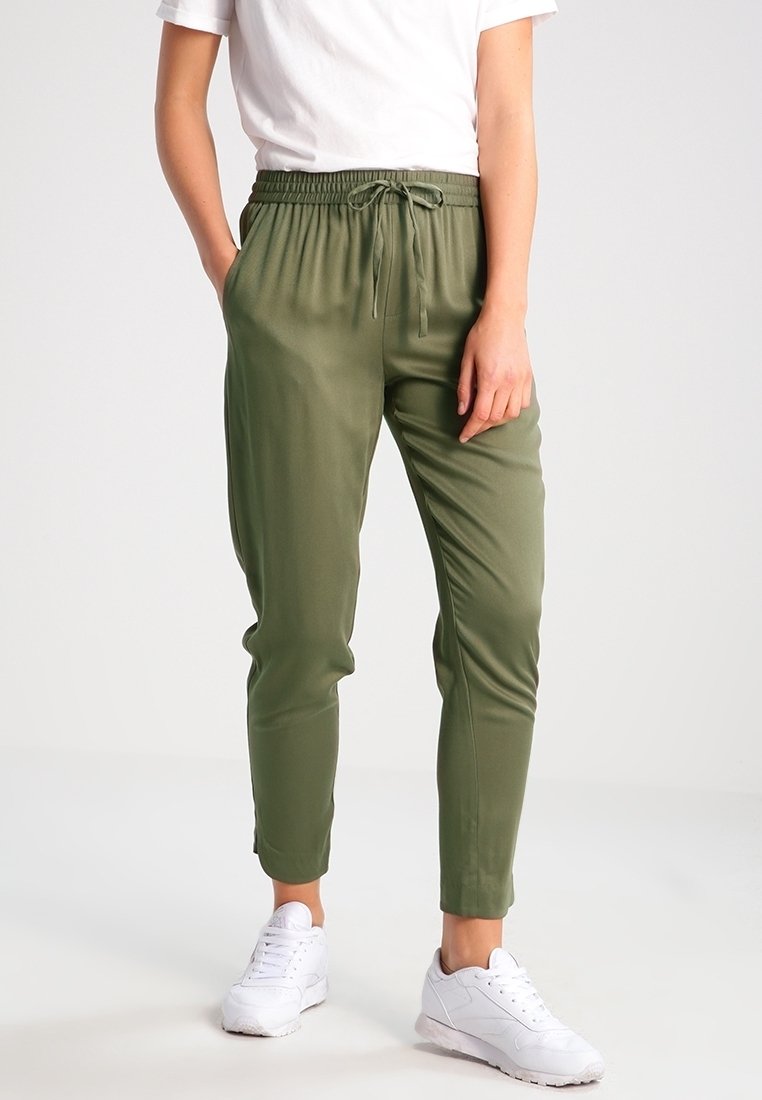 Personne portant un pantalon à cordon vert olive, des baskets blanches et une chemise blanche à manches courtes, debout avec une main dans la poche.