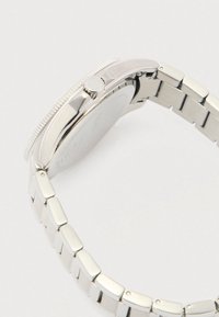 Sudraba metāla rokas pulkstenis ar apaļu korpusu, teksturētu rāmīti un plakanu saites dizainu. Ieklūst sānu kronis un gravēta aizmugure.