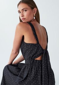 Žena so svetlou pokožkou a dlhými vlasmi má na sebe čierne šaty bez rukávov s bielymi bodkami, odhaleným chrbtom a viazanou mašľou, pozerajúc sa cez svoje rameno.