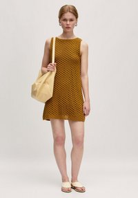 Ärmelloses Strickkleid in Senfgelb mit offenem Webmuster, kombiniert mit einer weichen beigefarbenen Tasche. Die Sandalen sind leicht und flach.