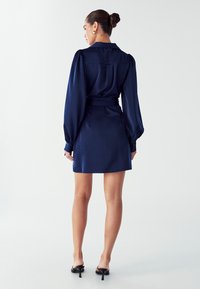 Námorníckomodrá saténová šaty s golierom, dlhými rukávmi s puffmi a viazaným pásom. Má krátku dĺžku a hladkú textúru.