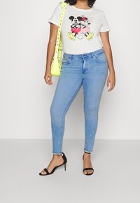 Camiseta blanca con gráfico negro y rosa de Mickey y Minnie, combinada con jeans skinnies azules claros y tacones negros con tiras. Bolso de mano verde neón.