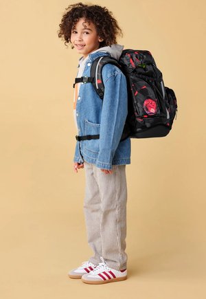 Lächendes Kind mit lockigem Haar, das gestreifte Hosen, Jeansjacke, weiße Turnschuhe und einen großen schwarzen und roten Rucksack trägt und seitlich steht.