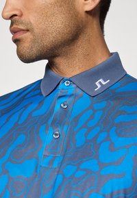 Homme portant un polo à motifs bleu et gris avec un col gris et un logo brodé blanc sur le col.