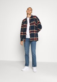 Flanellen overhemd met een marineblauw, rood en crème geruite patroon, met twee borstzakken en knoopsluiting. G gedragen over een wit t-shirt met blauwe jeans.