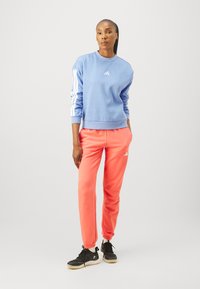 Ljusa blå sweatshirt med vit logga, accentuerad med vita ränder på ärmarna. Korallfärgade sweatpants med avsmalnande botten, som har vit logga. Svarta skor.