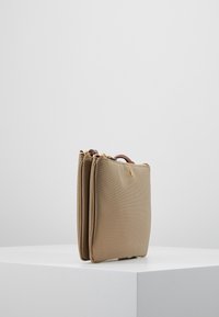 Borsa in tessuto beige con forma strutturata, dotata di un flap frontale, due zip superiori e un manico marrone. Accenti in hardware dorato visibili.