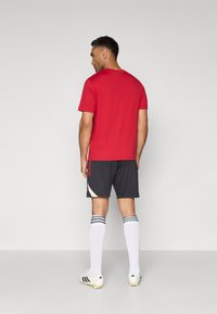 T-shirt rouge à manches courtes, assorti à un short noir à rayures, chaussettes blanches et chaussures de football avec des accents noirs et dorés.