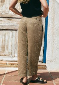 Hellbraune gemusterte Hosen mit entspannter Passform, geradem Bein, zwei Gesäßtaschen und Knopfdetails, kombiniert mit dunklen Sandalen.