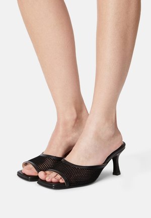 Woman's feet wearing black open-toe mesh high-heeled mules with square toes against a plain white background.
